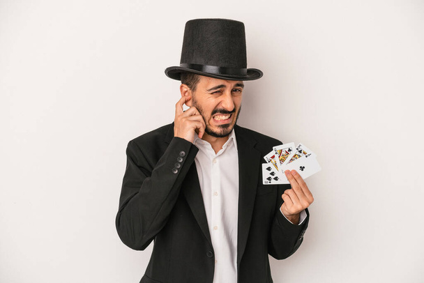 Νεαρός μάγος άνθρωπος κρατώντας μια μαγική κάρτα απομονώνονται σε λευκό φόντο καλύπτει τα αυτιά με τα χέρια. - Φωτογραφία, εικόνα