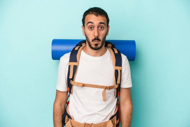 Young caucasian hiker man isolated on blue background shrugs shoulders and open eyes confused. - Photo, Image
