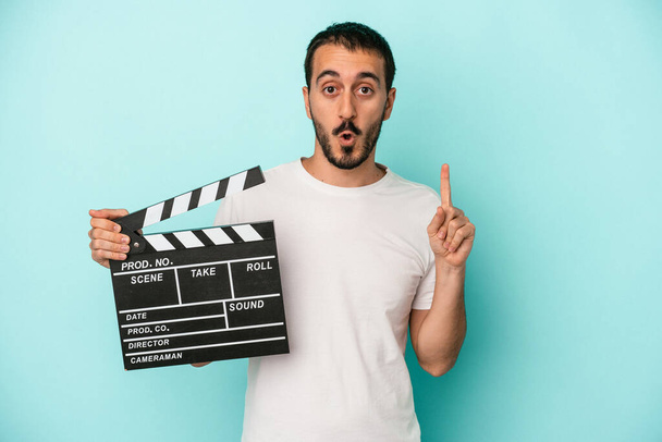 Jonge blanke acteur man houden clapperboard geïsoleerd op blauwe achtergrond met een aantal geweldige idee, concept van creativiteit. - Foto, afbeelding