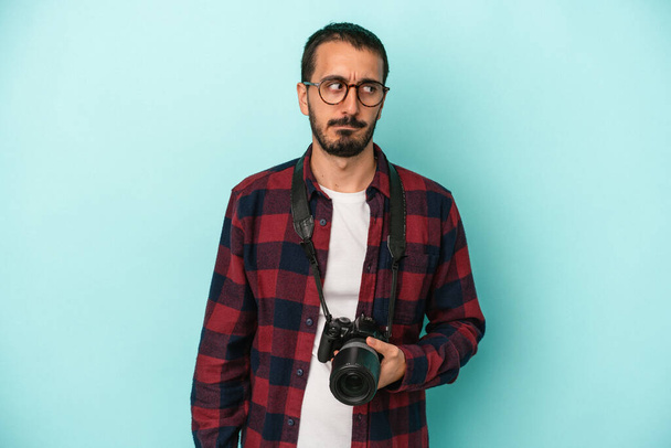 Молодой кавказский фотограф, изолированный на синем фоне, смущен, чувствует себя сомнительным и неуверенным. - Фото, изображение
