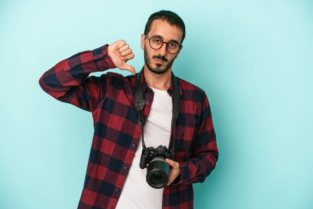 Junge kaukasische Fotograf Mann isoliert auf blauem Hintergrund zeigt eine ablehnende Geste, Daumen nach unten. Konzept der Uneinigkeit. - Foto, Bild