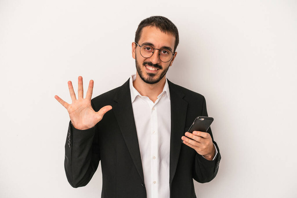 Young business man holding a mobile phone isolated on white background smiling cheerful showing number five with fingers. - Fotó, kép