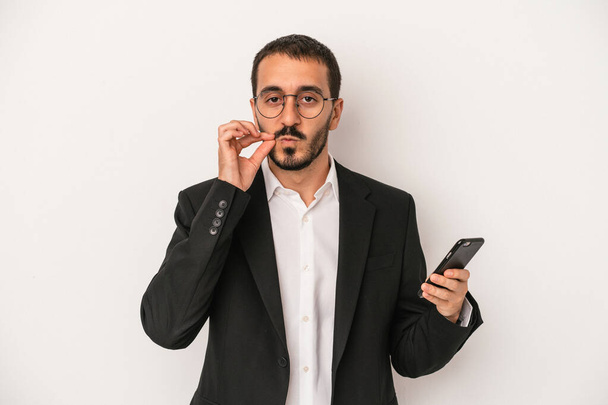 Young business man holding a mobile phone isolated on white background with fingers on lips keeping a secret. - Фото, изображение