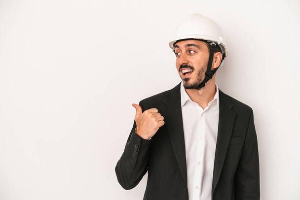 Young architect man wearing a construction helmet isolated on white background points with thumb finger away, laughing and carefree. - Photo, Image