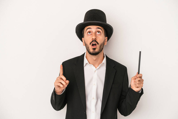 Mladý kouzelník muž drží hůlku izolované na bílém pozadí ukazuje vzhůru nohama s otevřenými ústy. - Fotografie, Obrázek
