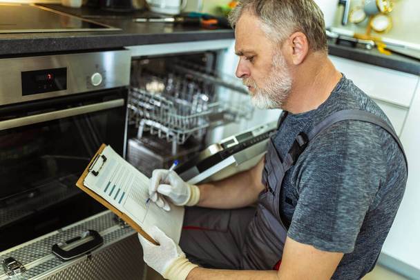 Técnico masculino sentado perto da máquina de lavar louça escrevendo na área de transferência na cozinha, close-up. - Foto, Imagem