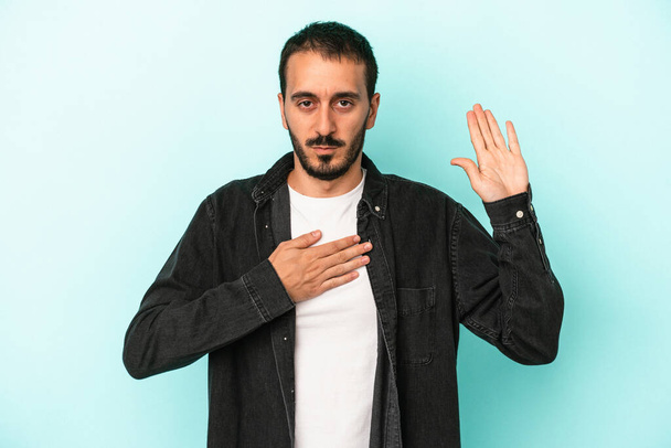 Молодой кавказский человек, изолированный на синем фоне, давая клятву, прикладывая руку к груди. - Фото, изображение