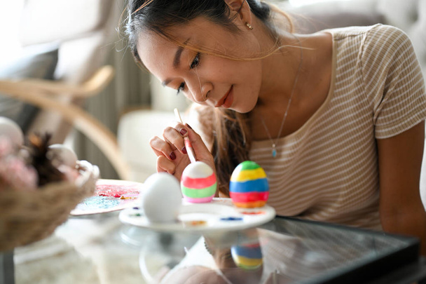 Relaxed and clam woman painting on an Easter egg with a colourful colour in her home living room. Happy Easter holiday concept. - Photo, image