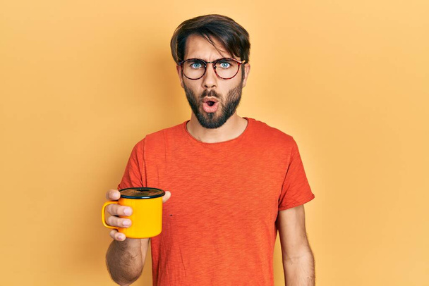 Joven hombre hispano bebiendo una taza de café asustado y sorprendido con la boca abierta para sorpresa, cara de incredulidad  - Foto, Imagen