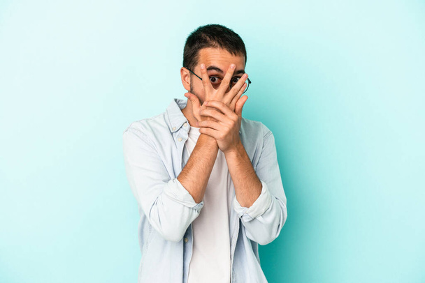 Νεαρός καυκάσιος άντρας απομονωμένος σε μπλε φόντο αναβοσβήνει μέσα από δάχτυλα φοβισμένος και νευρικός. - Φωτογραφία, εικόνα