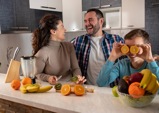 Μητέρα και πατέρας να στέκονται στην κουζίνα γελώντας με τον γιο τους που χαζολογάει κρατώντας πορτοκάλια στα μάτια. - Φωτογραφία, εικόνα