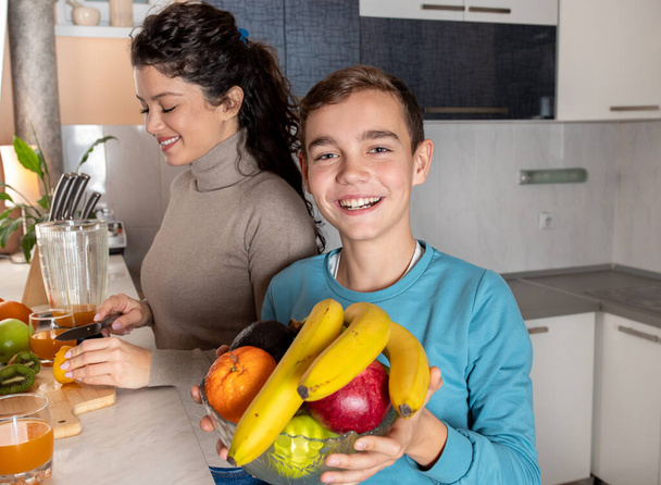 Sonriente niño sosteniendo un tazón de fruta en su mano mientras su madre cortaba naranjas y le hacía un jugo de naranja fresco - Foto, imagen