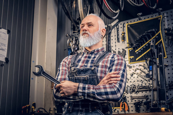 Літній ремонтник з сірою бородою тримає гайковий ключ у ремонтному магазині
 - Фото, зображення