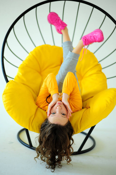 rocking chair a girl in a bright yellow sweatshirt sits on a juicy yellow chair and writhes faces different emotions on the white background of the studio teenager is having fun and she is smiling - Photo, Image
