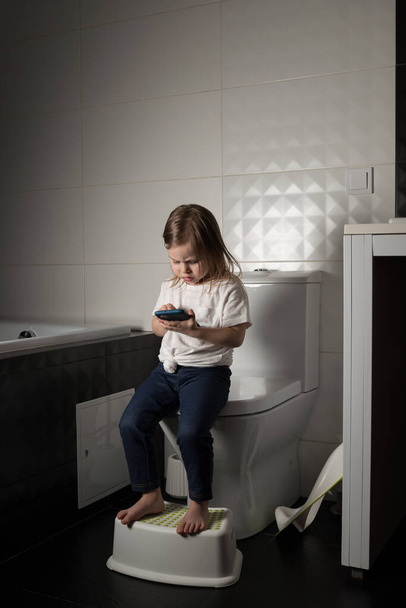A girl dressed in a blue jeance and white t-shirt playing with a mobile phone in the bathroom - Photo, Image