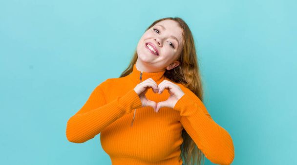 pretty red head woman smiling and feeling happy, cute, romantic and in love, making heart shape with both hands - Photo, Image