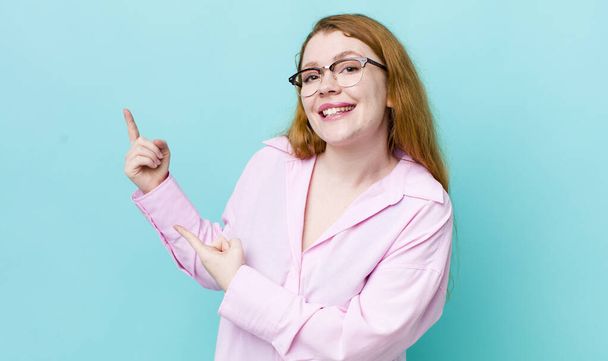 pretty red head woman smiling happily and pointing to side and upwards with both hands showing object in copy space - 写真・画像