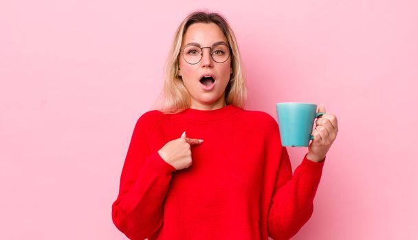 blonde pretty woman looking shocked and surprised with mouth wide open, pointing to self. coffee cup concept - Foto, immagini