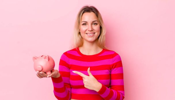 blonde pretty woman smiling cheerfully, feeling happy and pointing to the side. piggy bank concept - Foto, immagini