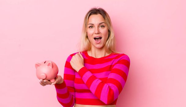blonde pretty woman feeling happy and facing a challenge or celebrating. piggy bank concept - Foto, immagini