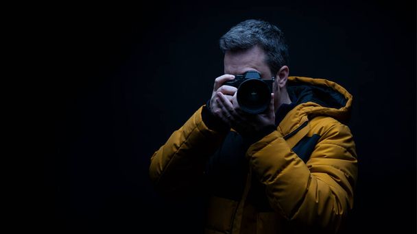 Προσωπογραφία φωτογράφου με κίτρινο χειμωνιάτικο παλτό που βγάζει φωτογραφία με μαύρο φόντο στο στούντιο - Φωτογραφία, εικόνα