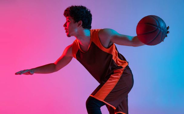 Young energetic man playing basketball isolated on gradient pink blue studio background in neon light. Youth, hobby, motion, activity, sport concepts. - Foto, Imagem