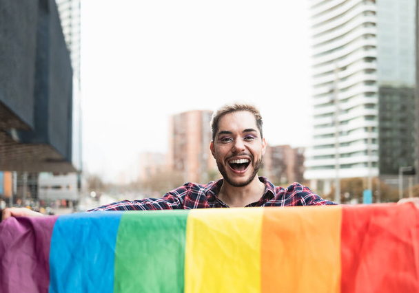 Glücklicher homosexueller Mann, der Spaß dabei hat, die Regenbogenfahne als Symbol der LGBTQ-Gemeinschaft zu halten - Foto, Bild