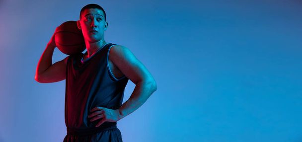 Portrait of young Korean man with basketball ball looking away isolated on blue studio background in neon light. Youth, hobby, sport concepts. - Foto, Imagem