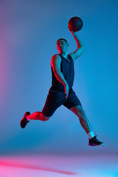 Junger, sportlicher Mann, Basketballspieler, der mit Ball auf blauem Studiohintergrund in Neonlicht springt. Jugend, Hobby, Bewegung, Aktivität, Sportkonzepte. - Foto, Bild