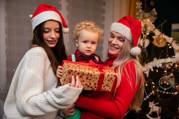 A mom and big sister give a gift to a two-year-old child at Christmas. - Fotó, kép