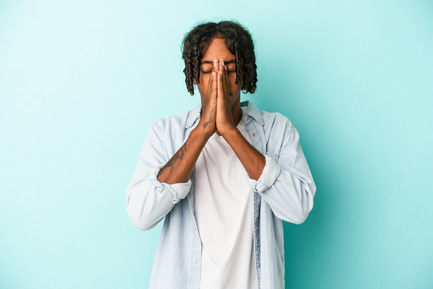 Joven afroamericano aislado sobre fondo azul cogido de la mano en orar cerca de la boca, se siente seguro. - Foto, Imagen