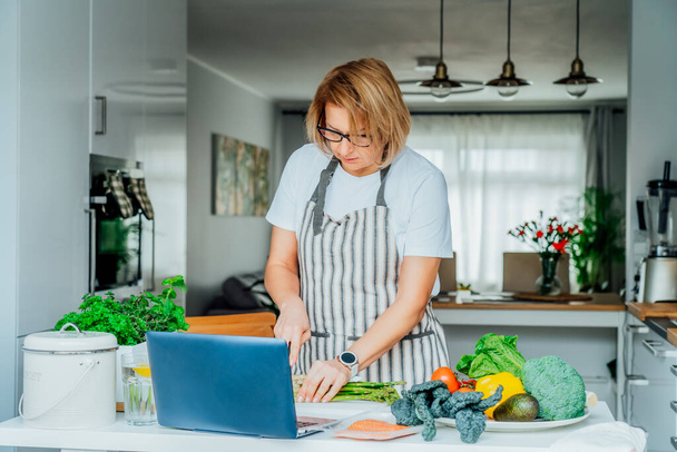 Middle age woman following a cooking tutorial video course on laptop while preparing meal in a kitchen. Woman cooking healthy dish, fish and vegetables on the table. Online recipe. Selective focus. - Foto, Bild