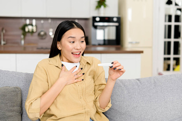 Surprised happy young woman sits on the couch at home with pregnancy test and celebrating result, rejoices to expected motherhood - Фото, изображение