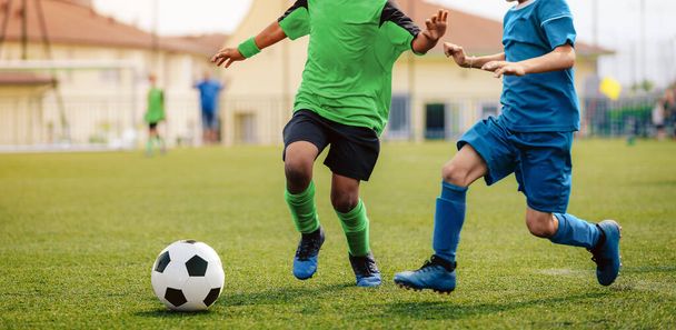 Two soccer players kicking soccer ball in a game. School boys play sports competition. Two multiracial kids playing football match. Kids in green and blue soccer jersy uniforms - Photo, Image