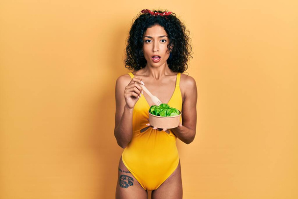 Joven chica latina con traje de baño comiendo ensalada en cara de shock, con aspecto escéptico y sarcástico, sorprendida con la boca abierta  - Foto, Imagen