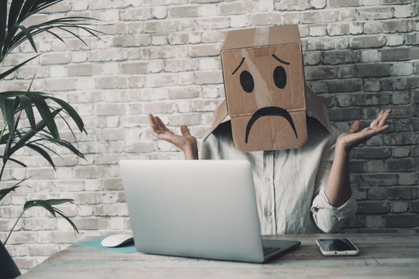 Modern worker with carton sad box on head open arms with desolation gesture in front of a laptop. Online crypto smart working business job activity. Concept of fail and lost work. Bad day office - Foto, Imagem