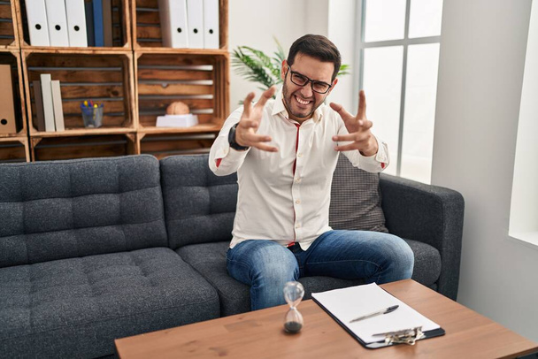 Junger hispanischer Mann mit Bart, der in der Beratungsstelle arbeitet, schreit frustriert vor Wut, Hände versuchen zu würgen, schreit verrückt  - Foto, Bild