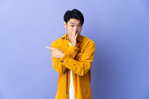 Молодий китайський чоловік, ізольований на пурпуровому фоні, вказує на бік, щоб представити продукт і прошепотів щось. - Фото, зображення