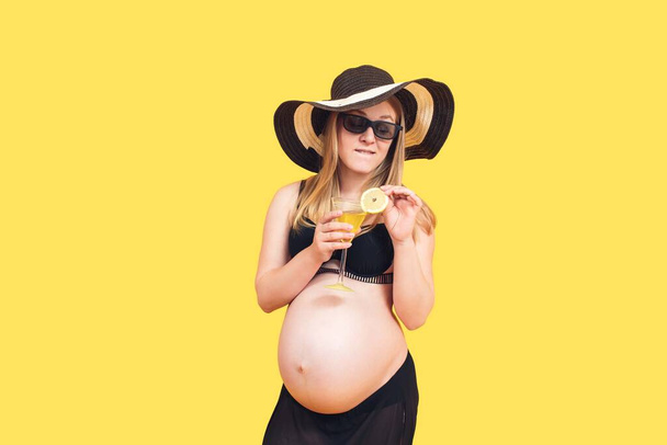 attractive pregnant woman on a yellow background holds a glass with a slice of lemon in her hands. - Fotoğraf, Görsel