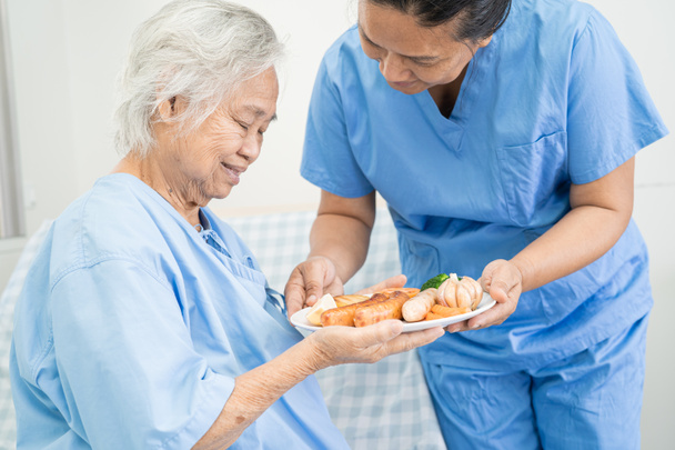 アジアの高齢女性高齢者の女性病院でベッドに座って空腹ながら希望と幸せと朝食と野菜の健康食品を食べる患者. - 写真・画像