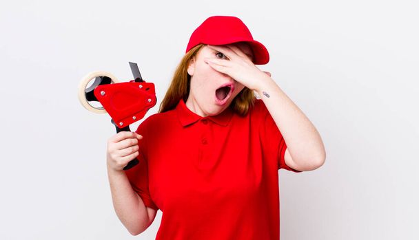 red head pretty woman looking shocked, scared or terrified, covering face with hand. packer employee concept - Foto, imagen