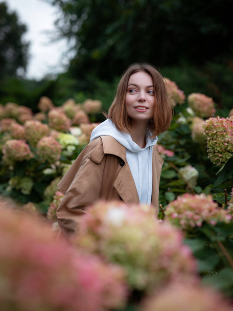 Beautiful woman in beautiful beige coat walk in autumn cloudy Moscow, in blooming hydrangeas. Street style. Travel to Russia concept. - Photo, image