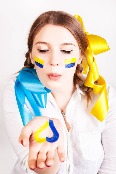 Молодая женщина с раскрашенным сердцем в ладони и украинским флагом посылает воздушный поцелуй на белом фоне. Мир и любовь. - Фото, изображение