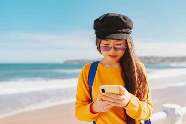 nuori aasialainen tyttö lasit ja baskeri konsultointi karttoja hänen älypuhelin kun sightseeing Euroopan kaupungissa. Kiinalainen nainen käyttää älypuhelinta - Valokuva, kuva