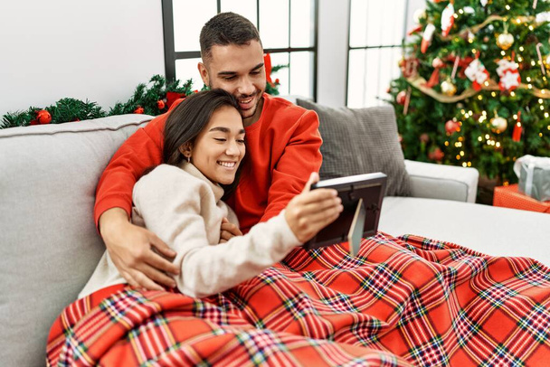 Joven pareja hispana sonriendo feliz y mirando la foto sentada en el sofá por el árbol de Navidad en casa. - Foto, Imagen
