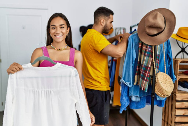 Joven pareja latina sonriendo feliz eligiendo ropa en la tienda de ropa. - Foto, imagen