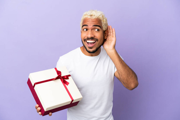 Young Colombian man holding a gift isolated on purple background listening to something by putting hand on the ear - Valokuva, kuva