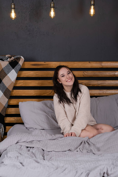 okouzlující mladá žena v pleteném béžovém svetru v posteli s šedým povlečením. moderní loft ložnice v hnědé a šedé barvě. - Fotografie, Obrázek