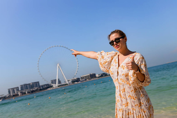 Счастливые женщины-туристки прогуливаются босиком по песчаному пляжу в районе аэропорта Дубая и любоваются панорамным видом на колесо обозрения Айн Феррис - Фото, изображение
