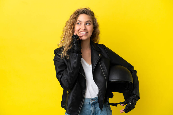 Fiatal szőke nő egy motorkerékpár sisak elszigetelt sárga háttér gondolkodás egy ötlet, miközben felnéz - Fotó, kép